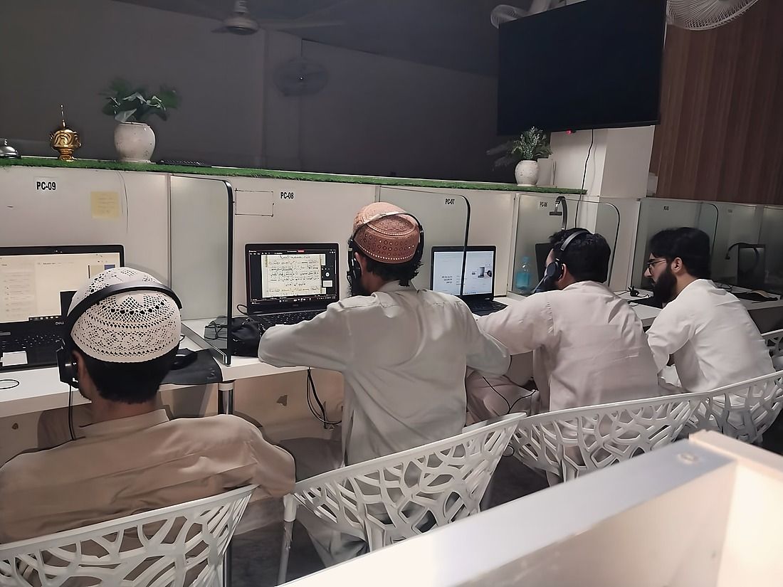 Teachers at their stations in the school’s teaching room, headsets on, a page of the Quran open on one of the screens