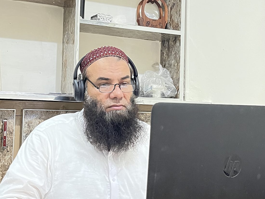Qari Muhammad Naveed Raja at his desk in the school, headset on, teaching a lesson online